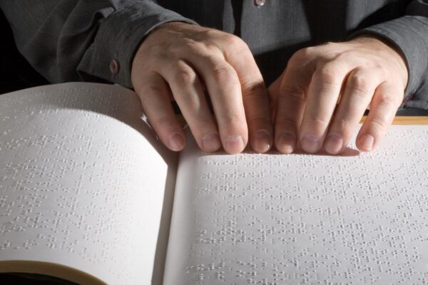 An image of a person from the chest down sitting at a desk guiding their hands across a book written in braille.