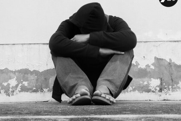 A homeless person sitting on the street with their head down in their lap and hood pulled over their head.